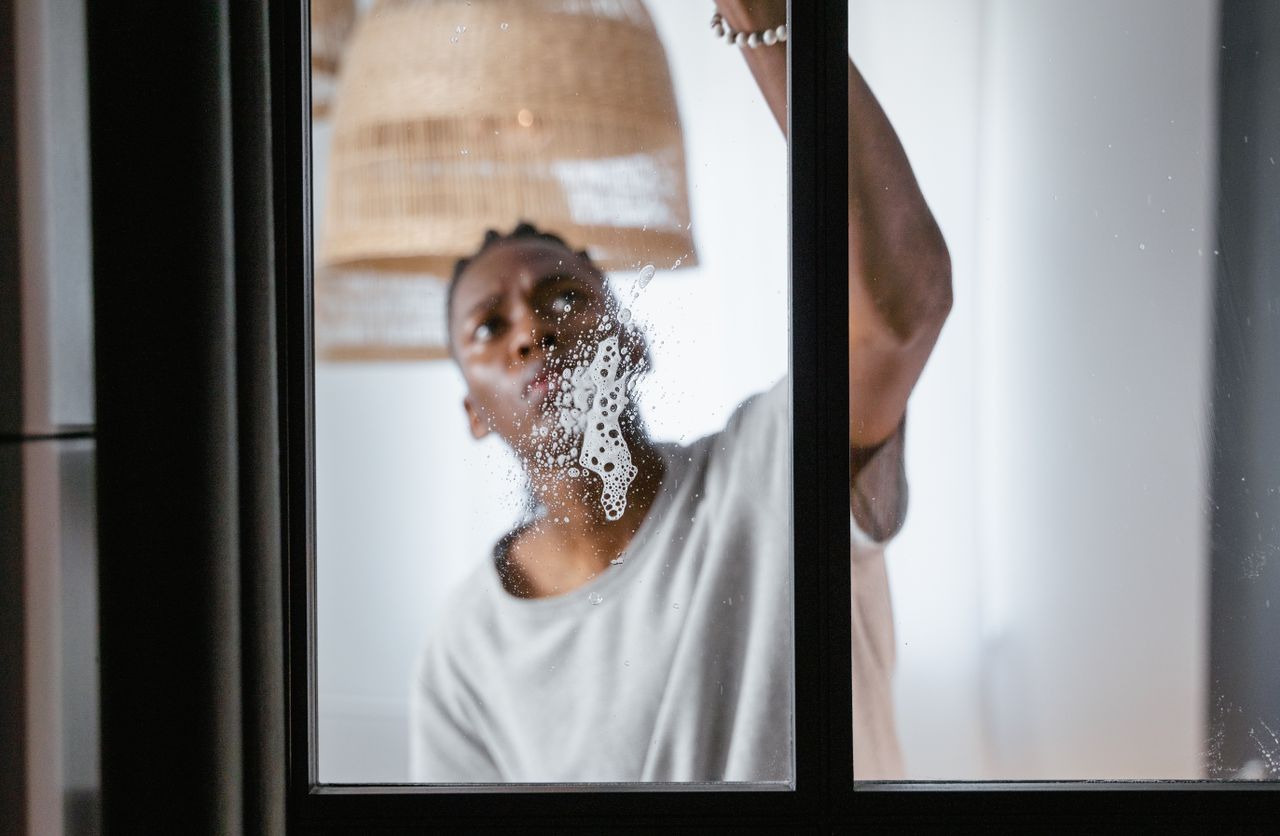 Un homme adulte nettoie une vitre à l’intérieur, dans le cadre des tâches ménagères et du maintien de la propreté.